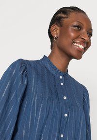 Camisa azul de botão com design pregueado, gola curta com babados e riscas verticais prateadas. Modelo a usar brincos circulares.