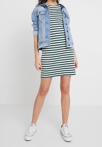 Pruhované zeleno-bílé šaty s kulatým výstřihem, doplněné světle modrou džínovou bundou a bílými teniskami. Neformální střih.