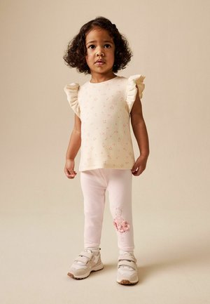 Young girl with curly hair wearing a cream ruffled sleeve top, pink leggings with a ballet girl design, and beige sneakers, standing on beige floor.