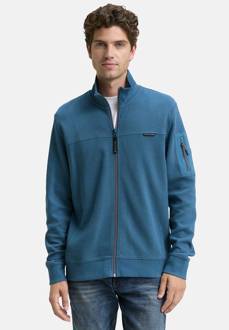 Blauwe fleece jas met rits en een gestructureerd patroon, staande kraag, contrasterende rits en borstzak, gedragen met spijkerbroek.