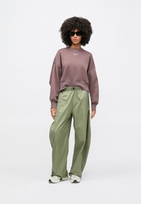 Άτομο με αφρο κόμμωση, φοράει μαύρα γυαλιά ηλίου, μωβ φούτερ Nike, φαρδιά ελιάς παντελόνια και λευκά αθλητικά παπούτσια σε λευκό φόντο.