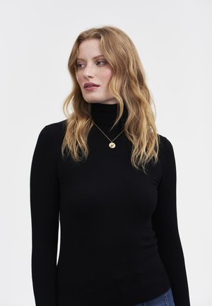 Eine Frau mit langen, welligen roten Haaren, die einen schwarzen Rollkragenpullover, eine blaue Jeans und eine goldene Anhänger-Halskette trägt. Sie schaut nach links vor einem weißen Hintergrund.