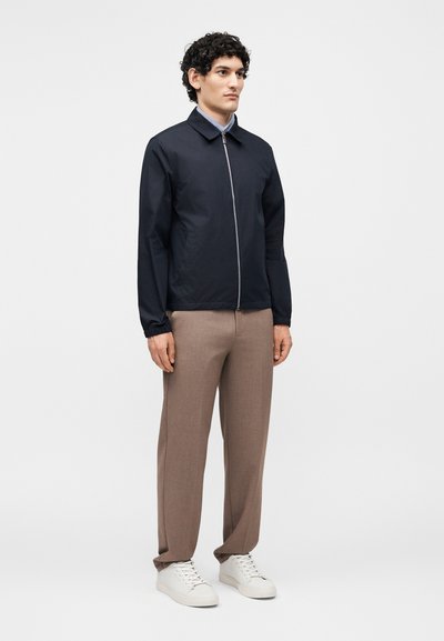 Άνδρας φοράει μαύρο μπουφάν με φερμουάρ, ανοιχτό μπλε πουκάμισο με γιακά, καφέ παντελόνι και λευκά αθλητικά παπούτσια, σε απλό λευκό φόντο.