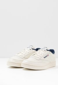 Zapatillas de cuero blancas de Reebok con detalles en azul marino, cordones planos, parte delantera perforada y suela de goma texturizada. Logo en el lateral y en la lengua.