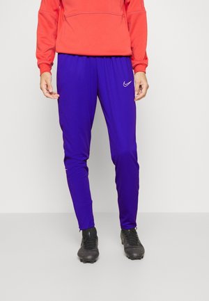 Osoba w jaskrawych fioletowych spodniach sportowych Nike, czarnych korkach oraz pomarańczowej bluzie, stojąca na jednolitym jasnym tle.