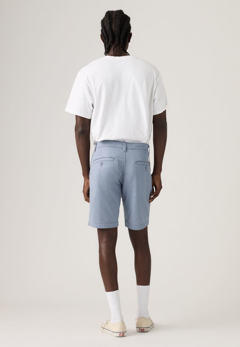 Homme aux cheveux tressés portant un t-shirt blanc, un short bleu clair avec des poches arrière, des chaussettes blanches et des baskets beige, debout de dos.