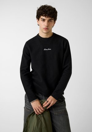 Jeune homme aux cheveux bouclés portant un pull noir Guess Jeans et un jean foncé, tenant une veste verte, debout devant un fond uni.