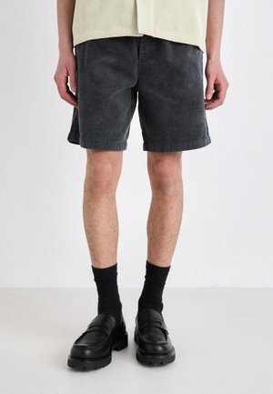 Shorts en velours côtelé gris avec une finition texturée, présentant une coupe décontractée et des ourlets roulés, associés à des mocassins noirs et des chaussettes de sport.