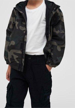 Niño con una chaqueta con cremallera de camuflaje en negro y gris sobre una camiseta blanca y pantalones cargo negros, con las manos en los bolsillos.