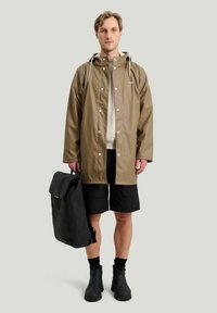 Brun vandtæt regnjakke med trykknaplukninger, parret med sorte shorts, sorte støvler og en sort rygsæk med rullet top.