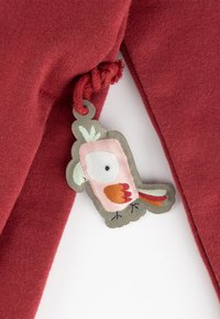 Roter Stoff mit glatter Textur, versehen mit einem dekorativen Vogelschild in Rosa, Grün und Orange mit gestickten Details und einer Schlaufe zur Befestigung.