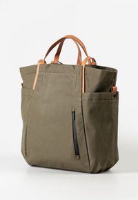 Olivgrüne Tragetasche mit beige Ledergriffen, Seitentaschen und einer vertikalen schwarzen Reißverschlusstasche vorne vor weißem Hintergrund.
