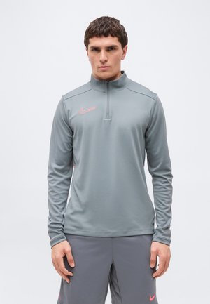 Un homme porte un haut de sport gris Nike à manches longues avec une fermeture éclair quart de zip et un short gris assorti avec des logos Nike roses, debout devant un fond blanc.