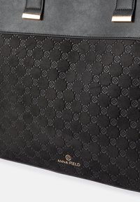 Black tote bag with a textured surface featuring an embossed pattern of circles and crosses. Gold logo detail at the bottom center.