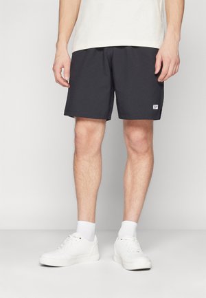 Schwarze Sportshorts mit strukturiertem Muster und Logoaufnäher, kombiniert mit weißen Turnschuhen und weißen Sportsocken.