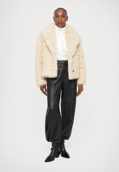 Giacca in finta pelliccia crema fuzzy con un ampio colletto, indossata sopra una camicia bianca e pantaloni in pelle nera, abbinata a stivaletti neri.