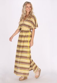 Abito lungo giallo, marrone e bianco con motivo a zigzag, maniche corte, vita stretta e spacchetti laterali, abbinato a sandali con tacco a zeppa intrecciati.