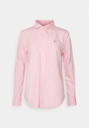 Chemise rose pâle à manches longues avec boutons sur le devant, col et petit logo brodé bleu sur le côté gauche de la poitrine, présentée sur un fond uni.