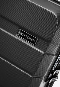 Valigia rigida in materiale nero texturizzato con scanalature diagonali, dotata di una placca logo argentata e di hardware in metallo per la cerniera.
