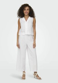 Completo con top bianco senza maniche e pantaloni a gamba larga, caratterizzato da dettagli in occhio di pernice all'orlo, realizzato in tessuto leggero, abbinato a sandali bianchi con cinturini.