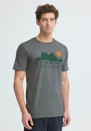 Junger Mann mit lockigem Haar trägt ein graues T-Shirt mit einer grünen Berggrafik und einer orangenen Sonne sowie dem Text "NORTH BEND Exploring the Wild" auf der Vorderseite.