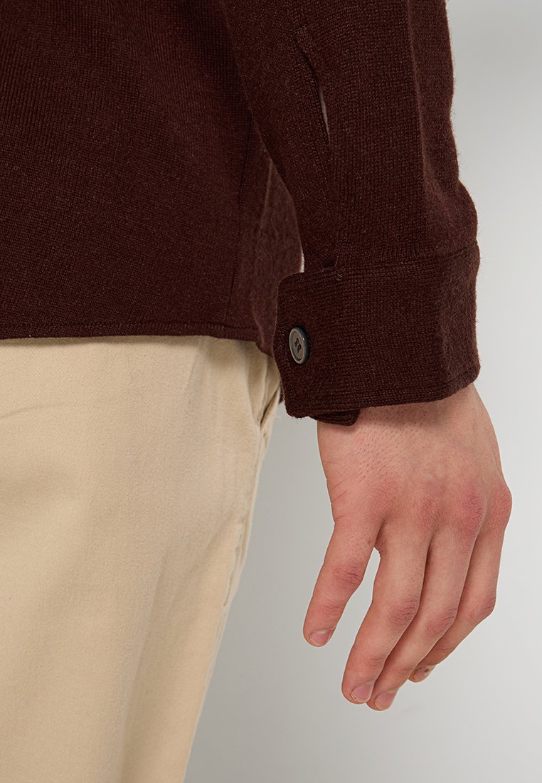 Bruine gebreide trui met een manchet met een knoopdetail, gedragen over beige broek, waarbij de textuur en pasvorm bij de pols zichtbaar zijn.