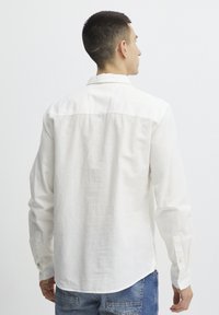 Camisa blanca de manga larga hecha de lino con un cuello abotonado y una textura suave. Presenta un dobladillo curvado y puños abotonados.