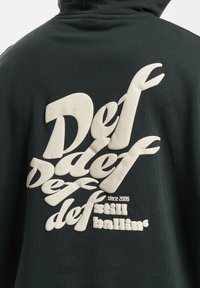 Mørkegrønn hettegenser med stor, hevet hvit skrift på ryggen, som viser ordene "Def" og "still ballin'" med en strukturert overflate.