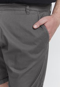 Pantalones cortos grises de textura suave, con bolsillos frontales, pasadores para cinturón y dobladillo doblado. Vista en primer plano con una mano en el bolsillo.