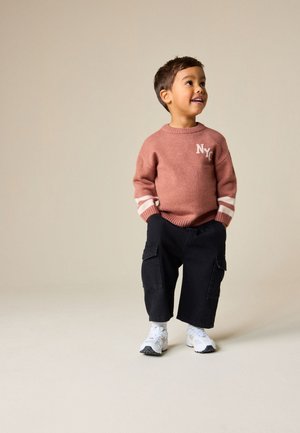 Jauns zēns smaida, valkājot rozā NYC džemperi, melnas kargo bikses un baltus sporta apavus, stāvot pret bēšu fonu.