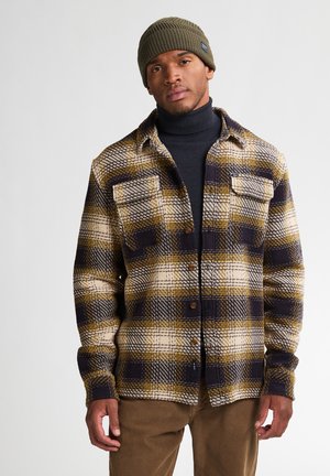 Chemise à carreaux marron, beige et bleu marine avec un devant à boutons, deux poches poitrine et des manches longues. Portée sur un col roulé noir et complétée par un bonnet vert.