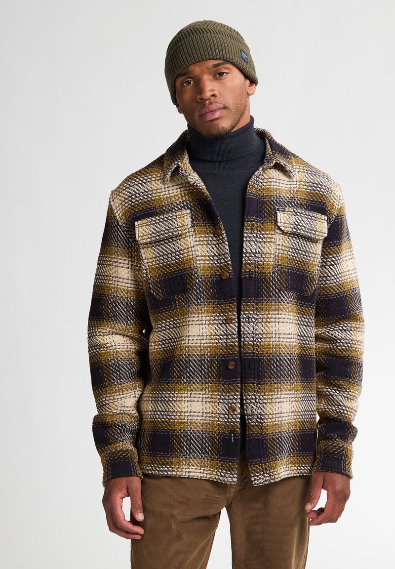 Geruite shirt in bruin, beige en marineblauw met knopen aan de voorkant, twee borstzakken en lange mouwen. Draagt over een zwarte turtleneck en aangevuld met een groene beanie.