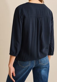Blusa blu navy con retro plissettato, maniche a tre quarti e scollatura rotonda; abbinata a jeans in denim blu con tasche posteriori.