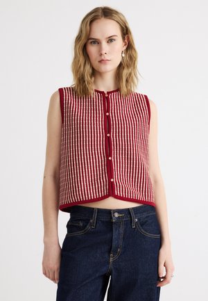 Jeune femme portant un gilet tricoté à rayures rouges et blanches sans manches, boutonné, avec un jean bleu foncé, sur un fond uni.