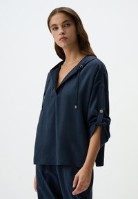 Pullover blu navy con scollo a V e cappuccio, dotato di lacci, maniche arrotolate e vestibilità morbida. Tessuto liscio e leggero.