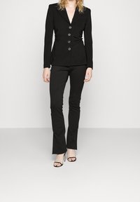 Černé na míru šité sako s hlubokým výstřihem do V, pěti knoflíky a strukturovaným střihem, doplněné úzkými černými kalhotami a černými sandály s otevřenou špičkou.