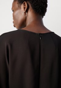 Zwarte kledingstuk met een gladde textuur en een wijde halslijn. Voorzien van een ritssluiting aan de achterkant en naaddetails voor een perfecte pasvorm.