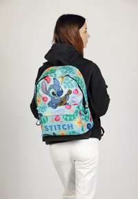 Mochila con un estampado tropical en azul y verde, con un personaje tocando la guitarra, el texto "STITCH" y patrones florales sobre un fondo claro.