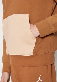 Personne portant un sweat-shirt marron et beige avec la main dans la poche avant et un pantalon marron assorti avec le logo Jumpman blanc.