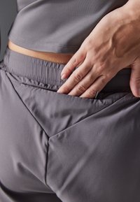 Harmaat urheiluhousut, joissa on kolmionmuotoinen taskumalli, joustava vyötärönauha ja sileä pinta. Käsi on asetettu taskulle.