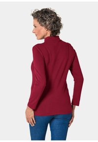 Bordeaux langemouwen top met hoge hals; gladde textuur, getailleerd ontwerp en effen kleur; gedragen met blauwe jeans.