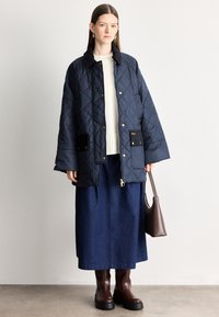 Tengerészkék steppelt külső ruházat magas gallérral, fekete zsebekkel és aranyszínű fém részletekkel. Krémszínű pulóverrel, farmer szoknyával és barna csizmával párosítva.