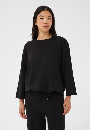 Femme aux longs cheveux foncés portant un haut noir ample à manches longues et un pantalon noir assorti sur un fond clair uni.