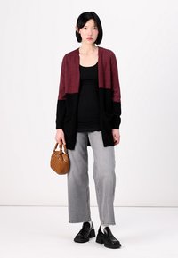 Cárdigan burdeos y negro con frente abierto, mangas largas, dos bolsillos; combinado con una camiseta de tirantes negra, jeans de pierna ancha grises y zapatos negros.