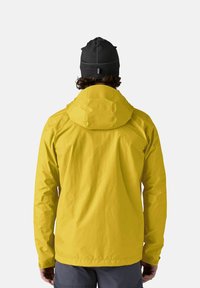 Chaqueta amarilla impermeable con capucha, hecha de tela suave, con mangas largas y puños elásticos, llevada con un gorro negro.