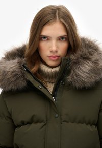 Chaqueta acolchada verde con capucha de pelo, con cierre de cremallera y detalles de botones. Debajo, se puede ver un grueso suéter de punto beige.
