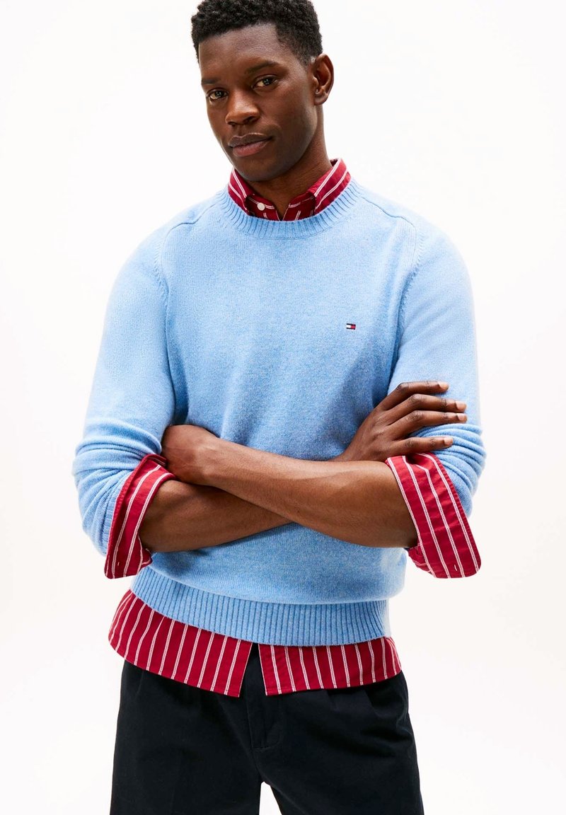 Maglione azzurro chiaro lavorato a maglia con orlo a coste, indossato sopra una camicia a righe rosse e bianche. Presenta un piccolo logo sul petto del maglione.