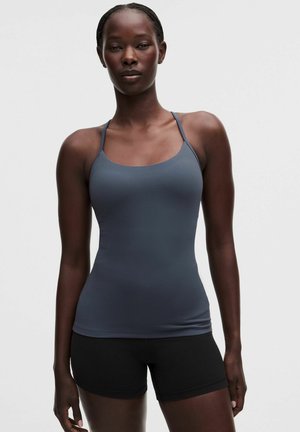 Kvinde iført en tætsiddende grå tanktop med tynde stropper og sorte shorts, der står mod en ensfarvet lys baggrund.
