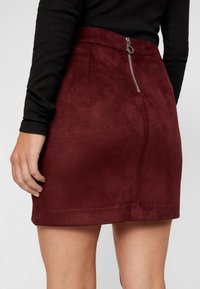 Mini-jupe en velours bordeaux avec une texture lisse, dotée d'une fermeture éclair au dos et sans poches visibles. Design simple et ajusté.