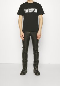 Svart bomull t-shirt med "THE KOOPLES" logotyp i vitt, kombinerad med svarta slim-fit läderbyxor och svarta snörskor.
